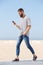 Full length happy man in earphones walking on beach with mobile phone