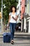 Full length laughing traveler walking on street with suitcase and smart phone