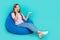 Full length photo of cute funny lady wear t-shirt sitting bean bag talking device empty space  turquoise color
