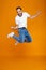 Full length photo of unshaved guy in t-shirt and jeans jumping and having fun, isolated over yellow background