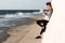 Full-length photo of young slim sport woman, using mobile phone, standing near white wall, seaside outdoor