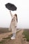 Full length portrait of the attractive brunette girl in white coat that stands among the dirt road, raised umbrella in