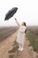 Full length portrait of the attractive brunette girl in white coat that stands among the dirt road, raised umbrella in