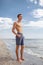 A full-length portrait of a boy with a muscular torso stands on a seashore on a natural blurred background.