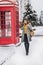 Full-length portrait of busy smiling woman talking on smartphone, walks by call-box. Outdoor photo of wonderful european