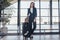 Full-length portrait of confident young business traveler wearing formal suit standing with heavy roll-aboard suitcase