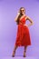 Full length portrait of content pretty woman 20s wearing red dress smiling at camera, standing isolated over violet background