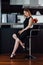 Full-length portrait of elegant woman with fair hair wearing black dress and high heels sitting on a bar chair in