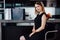 Full-length portrait of elegant woman with fair hair wearing black dress and high heels sitting on a bar chair in