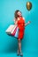 Full length portrait of a happy excited woman in red dress standing and holding colorful shopping bags and heart golden balloon is