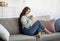 Full length portrait of millennial Indian lady drinking coffee, resting on sofa, smiling at camera, indoors