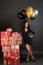 Full length portrait of a smiling stylish woman leaning on a pile of gift boxes and hiding balloons behind back, wears sparkly