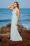 Full length portrait of traveller woman in blue chiffon dress enjoys her tropical beach vacation standing on a rock