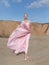 Full length portrait of a young blonde high girl in flying pink long dress poses in sand on sunny summer day