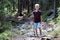 Full-length portrait of young child boy with hikers backpack and stick traveling alone through lit by bright sun mountain dense pi