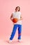 Full length portrait of young man dressed in retro sport uniform and holding basketball ball against pastel pink