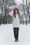 Full-length portrait of young woman in winter clothes who stands and adjusts her hat in snow-kept park. Vertical frame