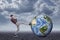 Full length side view of young man in casual clothes kicking huge Earth globe lying on ground under gray cloudy sky.