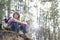 Full length of smiling male hiker looking away while sitting on cliff in forest