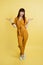 Full length studio shot of pretty young girl in yellow overalls and sneakers, posing on isolated pastel yellow