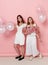 Full length view of a two of young friends, dressed in a white, holdings a balloons, over pink background.