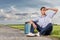 Full length of young man shielding eyes while sitting with petrol can by country road