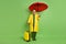 Full size photo of brunet optimistic guy with umbrella bag wear cloak boots isolated on green color background