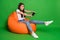 Full size photo of shocked girl sit orange bean chair play video game isolated over green shine color background