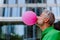 Fun portrait of happy energetic mature man inflating pink balloon in street, standing in front of skyscraper, copy space