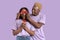 Funky black guy covering his girlfriend`s eyes with red hearts, surprising her on lilac studio background