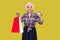 Funny aged woman in white hat and pink eyeglasses, in checkered shirt standing, holding bank credit card and shopping bags with