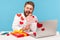 Funny bearded man talking video call sitting at workplace with laptop all covered with sticky hearts, showing pink heart,