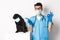 Funny black pug dog wearing medical mask, sitting near handsome veterinarian doctor showing okay sign, white background