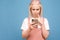 Funny blonde girl in cute clothes with a thoughtful face uses a smartphone on a blue background, the focus on the phone in the