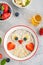 Funny bowl with oat porridge with owl faces made of fruits and berries on a gray concrete background. Food for kids idea, top view