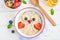 Funny bowl with oat porridge with owl faces made of fruits and berries on a white wooden background. Food for kids idea, top view