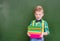 Funny boy holds a stack of heavy books near empty green chalkboard