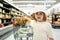 Funny boy with shopping cart full of fresh organic vegetables and fruits standing in grocery department of food store or