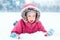 Funny Caucasian smiling girl in warm winter clothes pink jacket playing with snow. Cute child lying on ground during cold winter