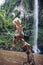 Funny child sit on snag under waterfall in tropical jungle