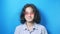 Funny curly-haired teenager in pink glasses looks into the camera and shows ok sign, on blue background