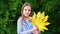 Funny cute girl teen with a large yellow autumn leaf in front of a green hedge