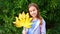 Funny cute girl teen with a large yellow autumn leaf in front of a green hedge