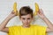 Funny face looking at camera. Banana as horns. Young boy in yellow t-shirt holding banana on his ears