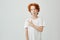 Funny ginger boy with freckles having curious expression and pointing a side with finger on white wall. Copy space for