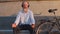 Funny gray-haired mustachioed bearded cyclist sits on city bench resting after trip. Active old man of retirement age