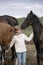 Funny happy child in a white sweater and jeans standing among horses foals on the farm smiling. Lifestyle portrait