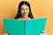 Funny hispanic woman reading a book doing crazy gesture with pencil over mouth like mustache, playful and positive on free time,