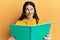 Funny hispanic woman reading a book doing crazy gesture with pencil over mouth like mustache, playful and positive on free time,