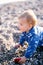 Funny kid in a blue blouse is crawling along a pebble beach, putting one hand on an apple and opening his mouth. Close